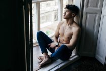 Продуманий молодий без сорочки чоловік сидить на підвіконні і дивиться геть — стокове фото