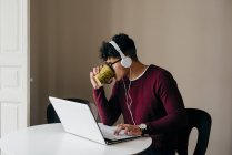 Молодой человек пьет кофе, печатая дома на ноутбуке — стоковое фото