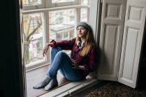 Cute blonde woman with headphones sitting at window and looking at camera — Stock Photo