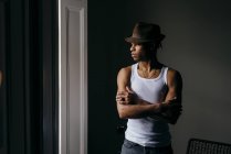 Stylish ethnic man in hat posing with crossed arms and looking at window — Stock Photo