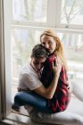 Junger Mann umarmt blonde Frau auf Fensterbank und zu Hause. — Stockfoto