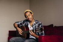 Веселий етнічний чоловік грає на гітарі і співає вдома — стокове фото