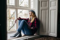 Улыбающаяся молодая женщина в наушниках на шее сидит у окна — стоковое фото