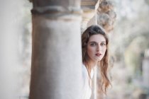 Hübsche junge Frau im weißen Kleid posiert an Säulen und blickt in die Kamera — Stockfoto