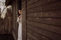 Young delicate brunette in white dress looking out of wooden doorway — Stock Photo
