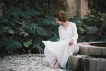 Brunette woman in white dress sitting at pond in park — Stock Photo