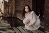 Jeune brune en robe blanche assise sur le seuil de la cabine en bois — Photo de stock