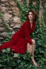 Чуттєва молода дівчина в червоній сукні сидить на плющах обійнятих скелях — стокове фото