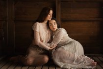 Young girls wearing old-fashioned elegant clothes and sitting in tender embrace on wood. — Stock Photo