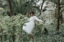 Mulher morena em vestido de luz branca girando em torno de plantas tropicais verdes . — Fotografia de Stock