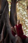 Chica rubia descalza en vestido rojo sentado en el tronco de enormes árboles en el bosque . - foto de stock