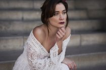 Young sensual brunette in white lace dress sitting on steps and looking away. — Stock Photo