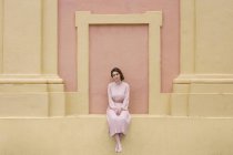 Frau in rosa Kleid sitzt an pinkfarbener Wand und blickt in Kamera — Stockfoto