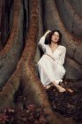 Brune rêveuse en robe blanche assise pieds nus dans les racines de vieil arbre avec les yeux fermés . — Photo de stock