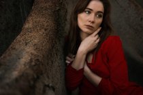 Sensual morena vestindo vestido vermelho e sentado por raízes — Fotografia de Stock