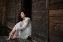 Young brunette in white dress sitting at wooden doorway with closed eyes — Stock Photo