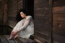 Jovem morena delicada em vestido branco sentado à porta de madeira — Fotografia de Stock