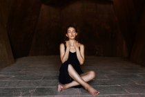 Giovane donna in abito nero seduta a piedi nudi e con gli occhi chiusi in camera di pietra . — Foto stock