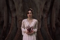 Mujer morena joven en vestido de encaje blanco y flores en el pelo sosteniendo follaje rojo oscuro - foto de stock