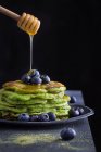 Honey pouring from wooden spoon to tasty green matcha pancakes with blueberries. — Stock Photo