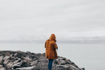 Anonymous person in coat standing on coast of gray rocks with misty water on background, Iceland. — Stock Photo