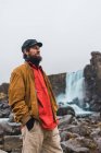 Bel homme barbu regardant loin tout en se tenant sur le fond de la belle cascade pendant le voyage à travers l'Islande. — Photo de stock