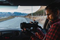 Bearded man in checkered shirt sitting on passenger seat of car and taking photos while traveling through Iceland. — Stock Photo