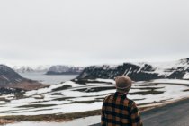 Back view of man in hat and shirt standing on background of snowy mountain range in lakes, Iceland. — Stock Photo