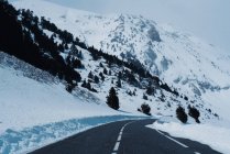 Perspective view of paved road running away among snowy slopes of snowy mountains with black trees. — Stock Photo