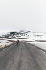 Back view of hipster guy riding skateboard on paved long road with snowy mountains on background in Iceland. — Stock Photo