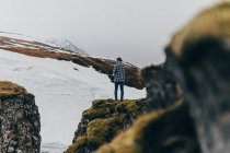 Rückenansicht eines lässigen Mannes, der auf grünem Felsen vor schneebedecktem Hochland im Nebel Islands steht. — Stockfoto
