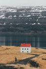 View of tourist walking on road towards small house placed alone on coast of cold lake in Iceland. — Stock Photo