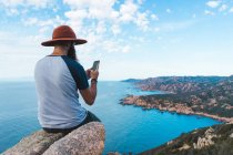 Man in hat sitting on rock at seaside and taking photo — Stock Photo