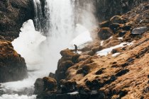 Vista a distancia del hombre de pie en el borde rocoso de la colina con salpicaduras de cascada en el fondo, Islandia. - foto de stock