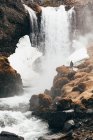 Vista a distanza dell'uomo in piedi sul bordo roccioso della collina con cascata schizzi sullo sfondo, Islanda. — Foto stock