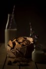 Kekse mit Schokolade im Papierbündel und eine Flasche Milch stehen auf einem Holztisch vor dunklem Hintergrund — Stockfoto