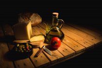 Натюрморт з помідорами, сиром, оливками, ножем, оливковою олією в мисці і хлібом, що стоїть на дерев'яному столі на темному фоні — стокове фото