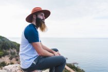 Uomo in cappello seduto sulla roccia al mare — Foto stock