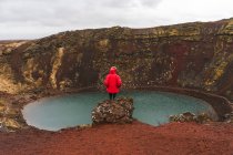 Back view of faceless man in warm clothes sitting on rim of open pit and looking at small lake while traveling through Iceland. — Fotografia de Stock