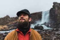 Handsome bearded man looking away while standing on background of beautiful waterfall during trip through Iceland. — Stock Photo