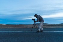 Vista lateral do homem em roupas quentes colocando câmera profissional no tripé enquanto tira fotos da natureza islandesa. — Fotografia de Stock