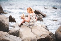 Redheaded woman sitting on rock at ocean — Stock Photo