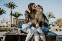 Jóvenes mujeres atractivas besándose y abrazándose mientras se sienta en la capucha del coche. - foto de stock