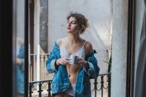 Жінка в джинсовій куртці тримає чашку на балконі — стокове фото