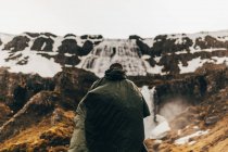 Vista posterior del hombre en impermeable verde de pie sobre el paisaje con montañas y cascada en Islandia. - foto de stock