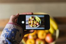 Human hand taking photo of fresh fruits in bowl with smartphone — Stock Photo