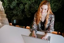 Cheerful young redhead woman sitting at laptop and typing at table — Stock Photo