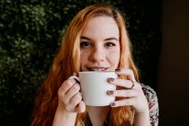 Усміхнена руда молода жінка з чашкою сидить проти куща — стокове фото