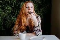 Fröhliche rothaarige hübsche Frau mit Tasse sitzt gegen Busch und isst Donut — Stockfoto