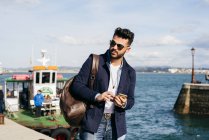 Человек с рюкзаком и смартфоном стоит в порту — стоковое фото